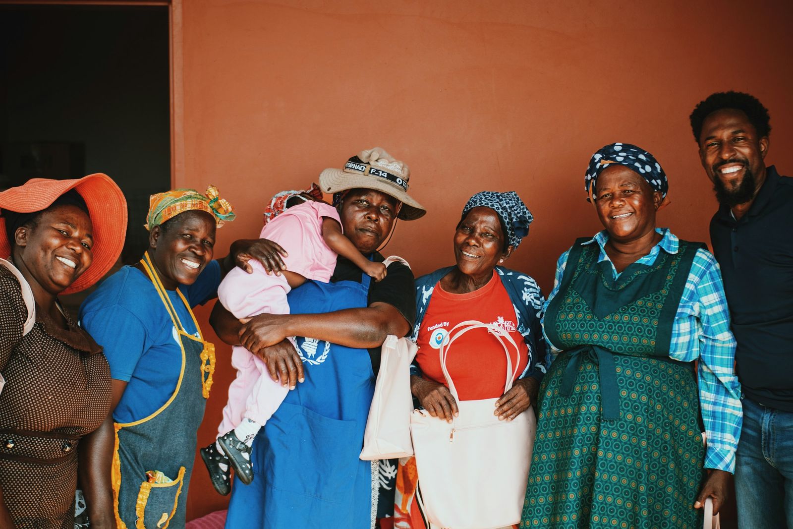 A smiling intergenerational group of Black women with an elder, a child, and a man, standing together against a terracotta wall